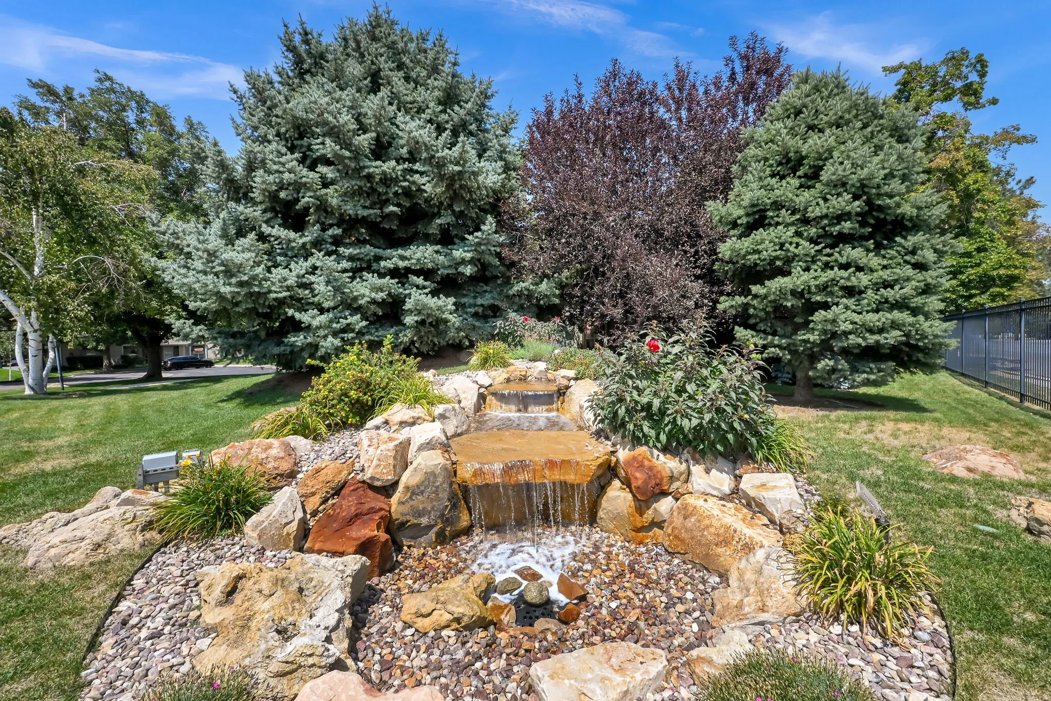 Mature landscaped grounds featuring a natural stone water feature with integrated seating, decorative river rock accents, and established shade trees throughout the property