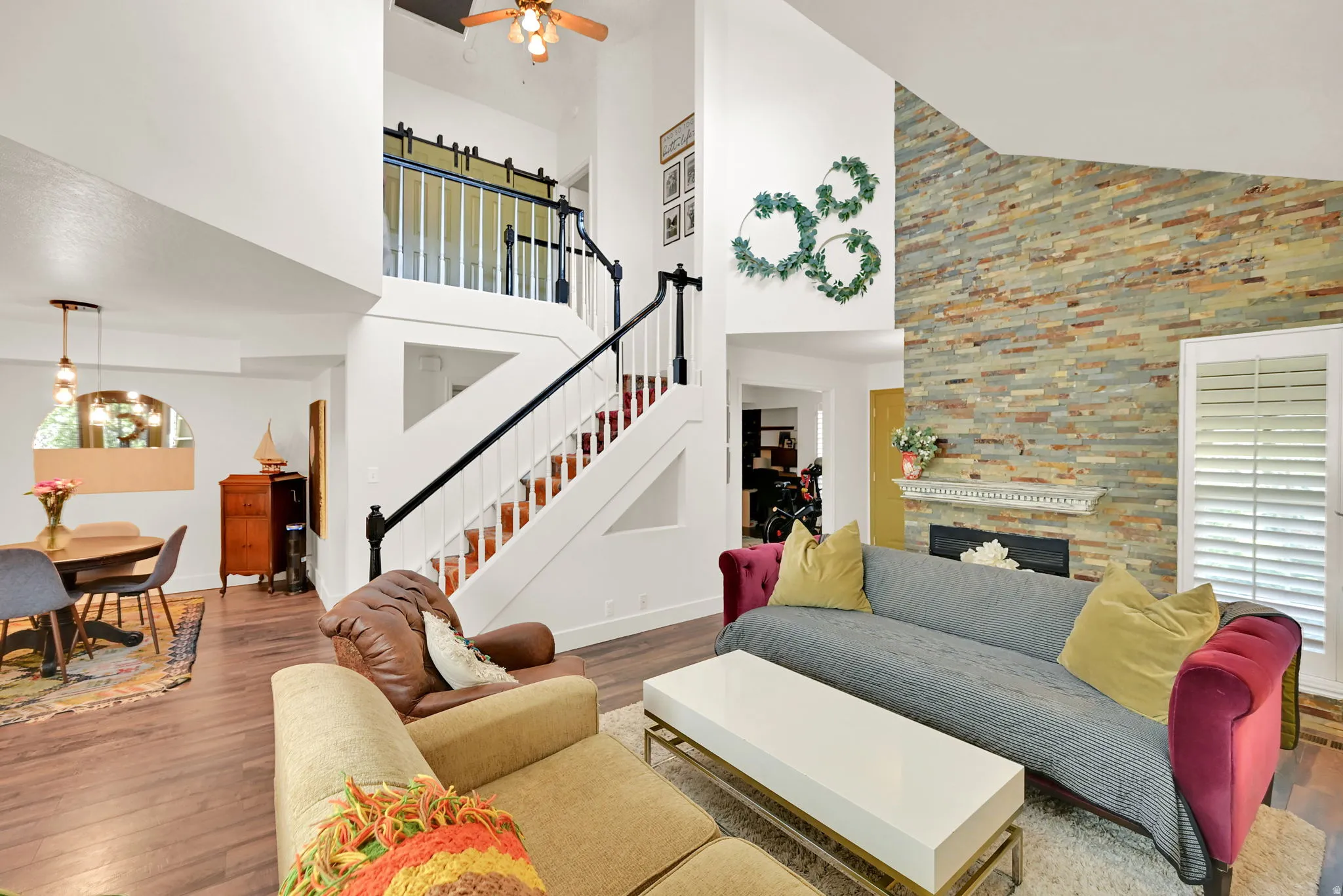 Open-concept living area showcasing soaring ceilings with ceiling fan, a dramatic open staircase with black metal railings and wood treads, wood-finish flooring throughout, and a striking stacked-stone accent wall