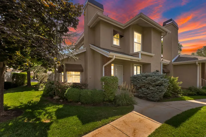Two-story residence featuring a taupe exterior with architectural detail, illuminated windows, and mature landscaping including evergreen shrubs and established trees