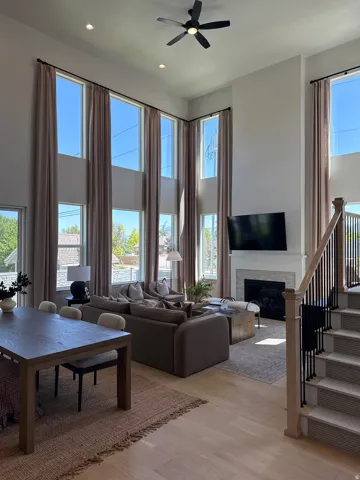 Two-story living area featuring extensive tall windows with neutral drapery, wood-finish flooring, a contemporary fireplace with a light-toned surround, and a staircase with black metal balusters