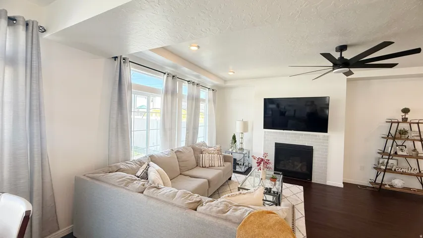 Bright, open living room with large multi-pane windows flooding the space with natural light, a wood-finish plank floor, a fireplace with a dark surround, a ceiling fan with multiple blades, and recessed lighting throughout