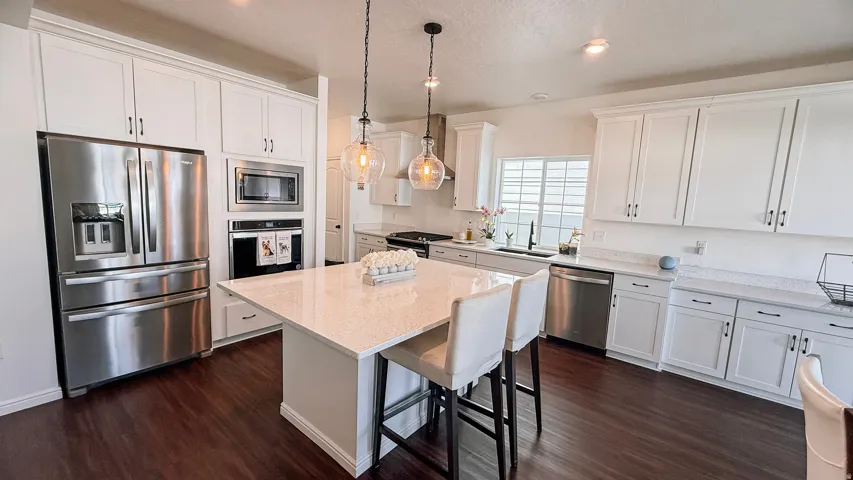 Spacious kitchen featuring white cabinetry, integrated stainless steel appliances, dark wood-finish flooring, a central island with a light-toned countertop, and pendant lighting fixtures