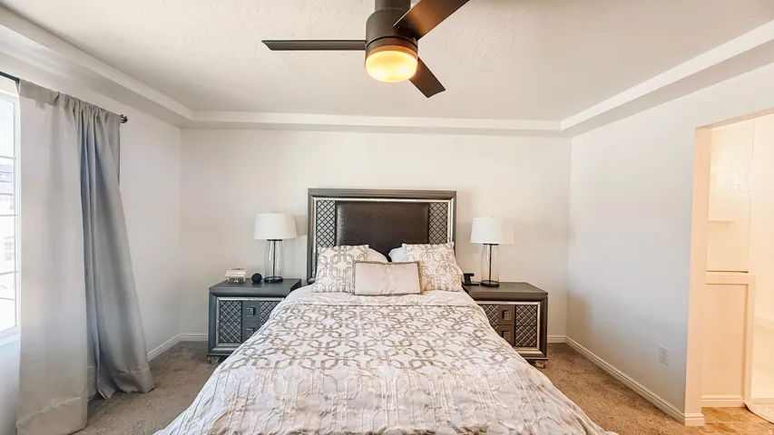 Tray ceiling, ceiling fan with integrated lighting, carpet flooring, window with trim, and a partial bathroom entrance