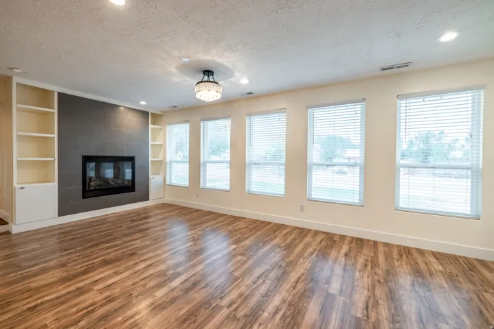 Spacious living room showcasing hardwood flooring, a gas fireplace with dark surround and built-in shelving, recessed lighting, and expansive multi-pane windows with horizontal blinds providing natural light throughout the space
