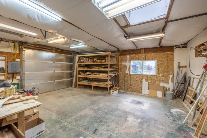Spacious shop featuring dual skylights for natural illumination, concrete flooring, wall-mounted shelving with wood-finish construction, a service garage door, and ample room for storage and projects