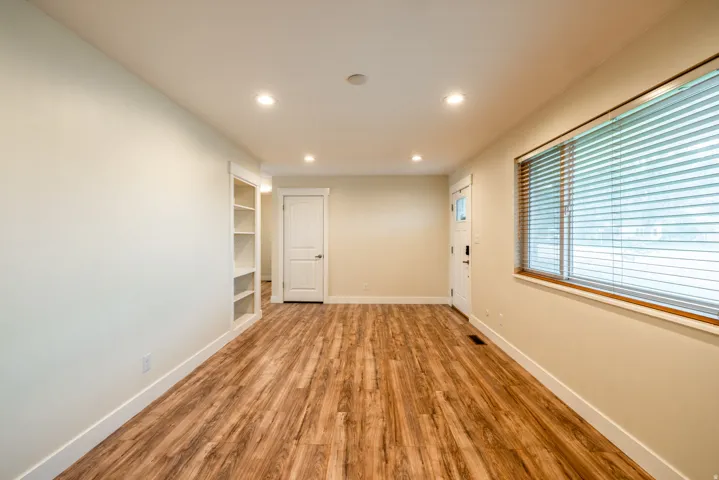Spacious room featuring newly installed wood-finish flooring, recessed ceiling lighting, built-in shelving along the wall, and large windows with horizontal blinds providing natural light and privacy