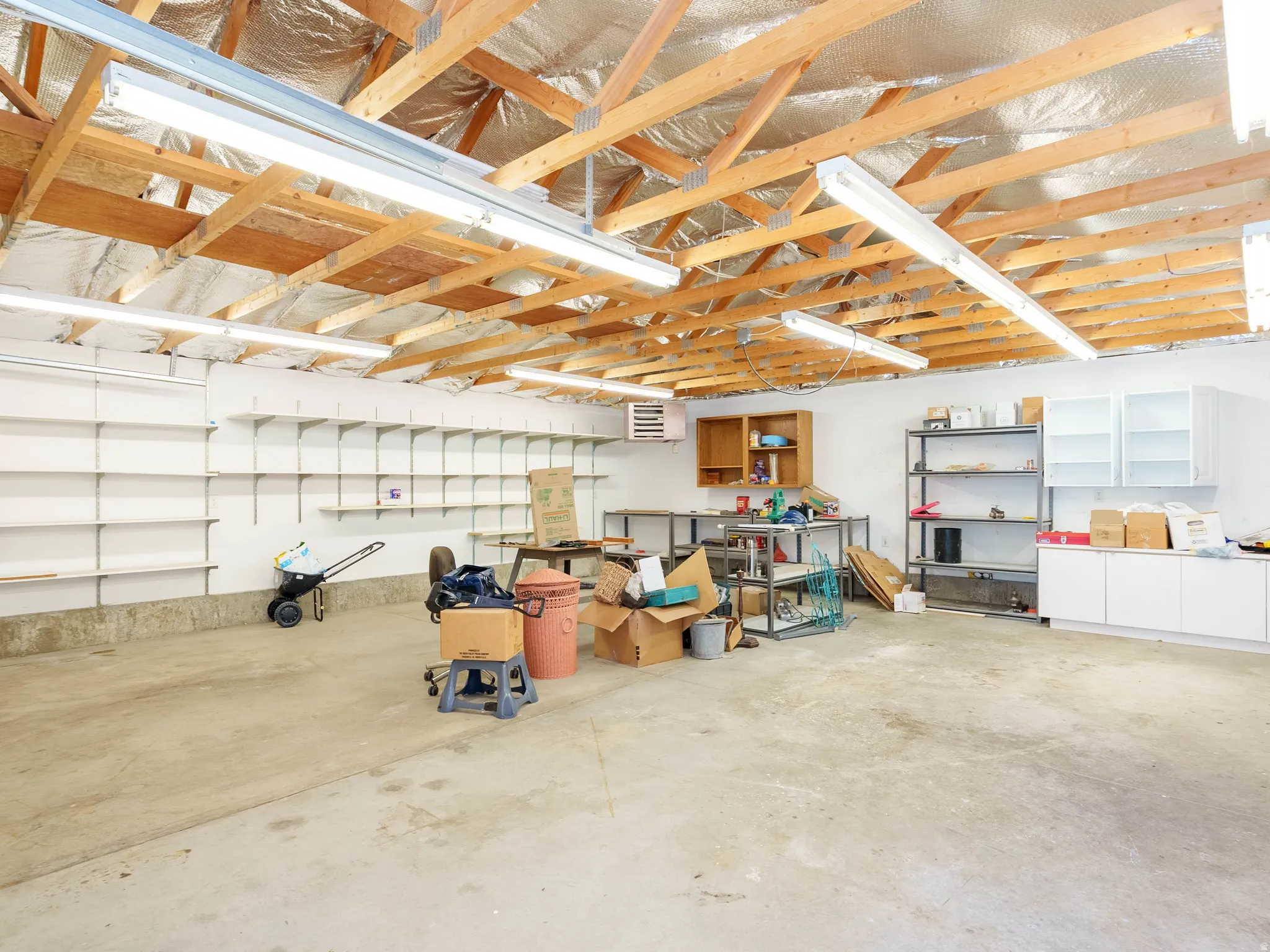 Exposed wood beam ceiling with reflective insulation, fluorescent lighting fixtures, concrete flooring, extensive wall-mounted shelving system, and white cabinetry