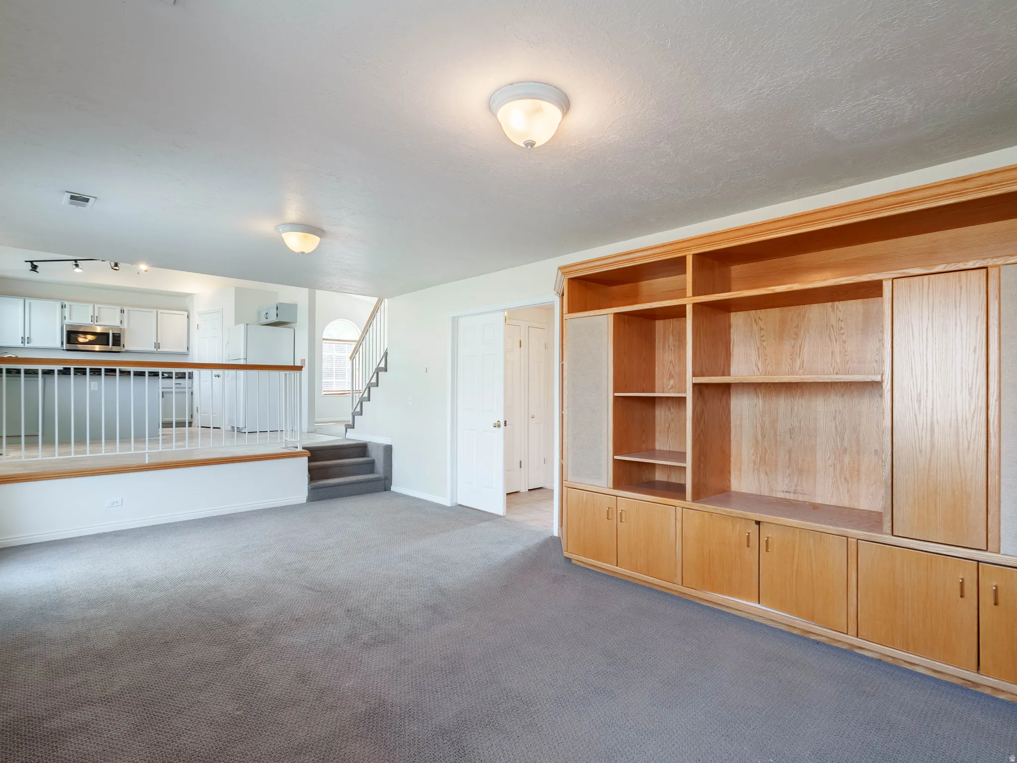 Built-in wood-finish cabinetry, gray carpeting, white kitchen cabinetry with track lighting, and a staircase with a white railing