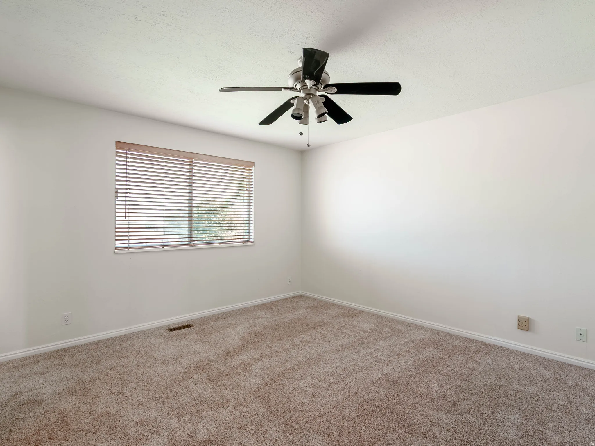 Bright, light-filled bedroom featuring neutral carpet flooring, large window with horizontal blinds providing natural light, and ceiling-mounted fan with integrated lighting fixture