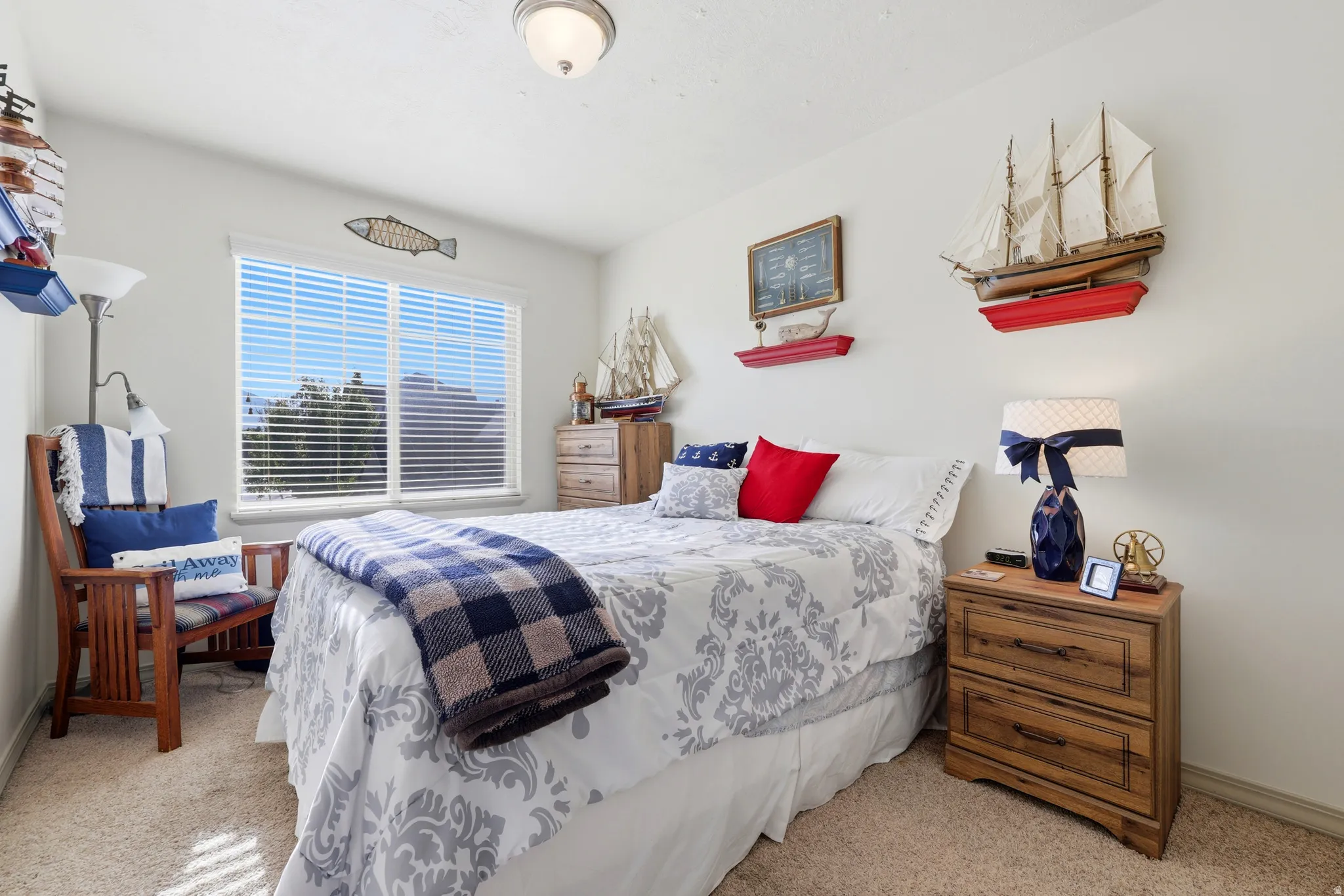 Recessed window with horizontal blinds, light-colored walls, ceiling-mounted light fixture, light-toned carpet flooring, and wood-finish nightstand with drawer storage