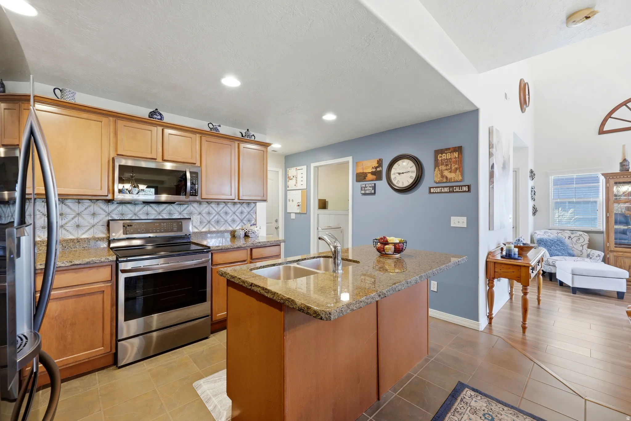 Kitchen featuring wood cabinetry, stainless steel appliances, a tiled backsplash, and an island with a built-in sink and granite countertop