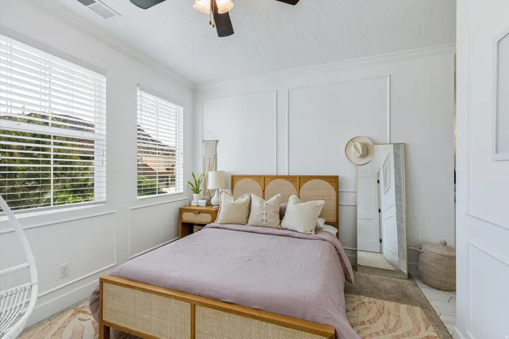 Bright bedroom offering plantation shutters, wood-frame bed with rattan panel headboard, coordinated bedding in soft neutrals, natural wood nightstands, ceiling fan with integrated lighting, and neutral area rug over wood-finish flooring