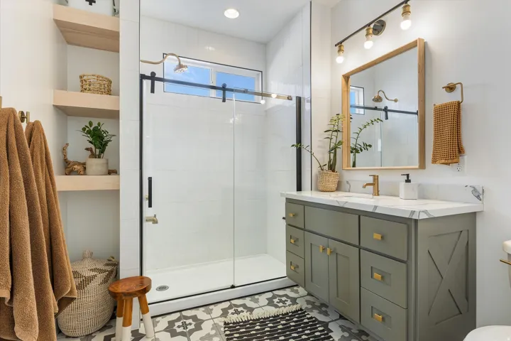 Spa-inspired primary bathroom featuring a frameless glass walk-in shower enclosure with matte black hardware, a gray shaker-style vanity with brass pulls and white countertop, large framed mirror with integrated lighting, and geometric patterned tile floo