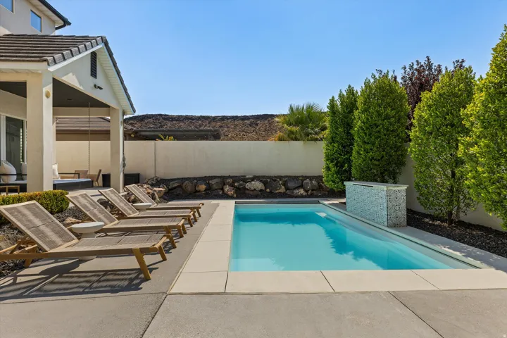 Resort-style backyard featuring a sparkling salt-water pool with clean decking, lounge seating areas, mature privacy hedging, and a modern home exterior with covered patio space overlooking the grounds