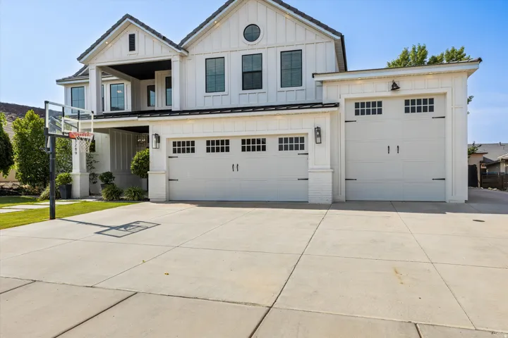Modern farmhouse exterior showcasing a three-car garage with paneled doors, expansive concrete driveway, crisp white siding with dark trim accents, gabled roofline with architectural detailing, and manicured lawn with mature landscaping