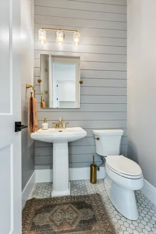 Elegant powder room featuring shiplap accent wall, pedestal sink with brass fixtures, hexagonal tile flooring, recessed medicine cabinet with framed mirror, and three-light vanity sconce above the sink