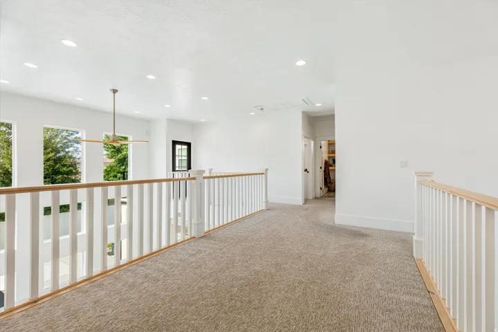 Spacious upper-level hallway featuring neutral carpet flooring, recessed ceiling lighting, white railing with wood-finish top rail, and abundant natural light from large windows