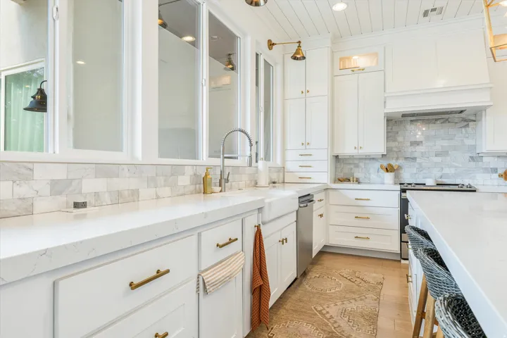 White shaker cabinetry with gold-tone hardware, wood-finish flooring, white countertop, farmhouse sink, and marble-look backsplash