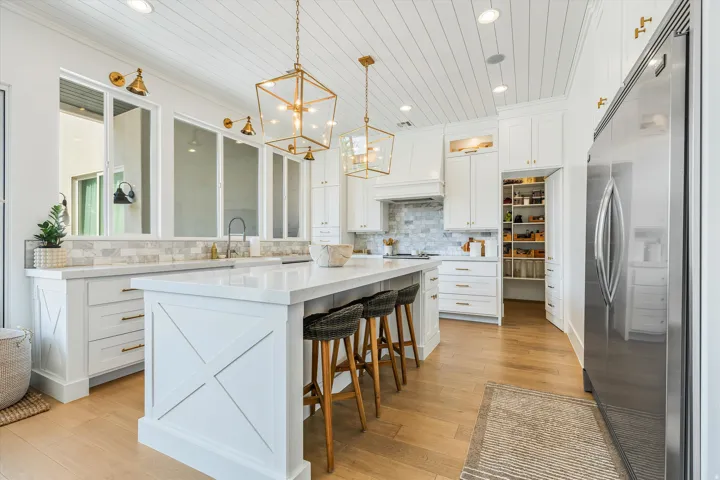 Gourmet kitchen featuring custom white cabinetry, a large waterfall-edge island with seating for multiple, wood-finish flooring, stainless steel appliances, geometric pendant lighting, and marble countertops with coordinating backsplash