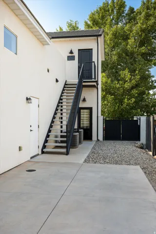 Modern architectural exterior featuring a clean white facade with black metal staircase and landing system, polished concrete patio, gravel hardscaping, mature landscaping with lush greenery, and black-framed entry doors with contemporary hardware