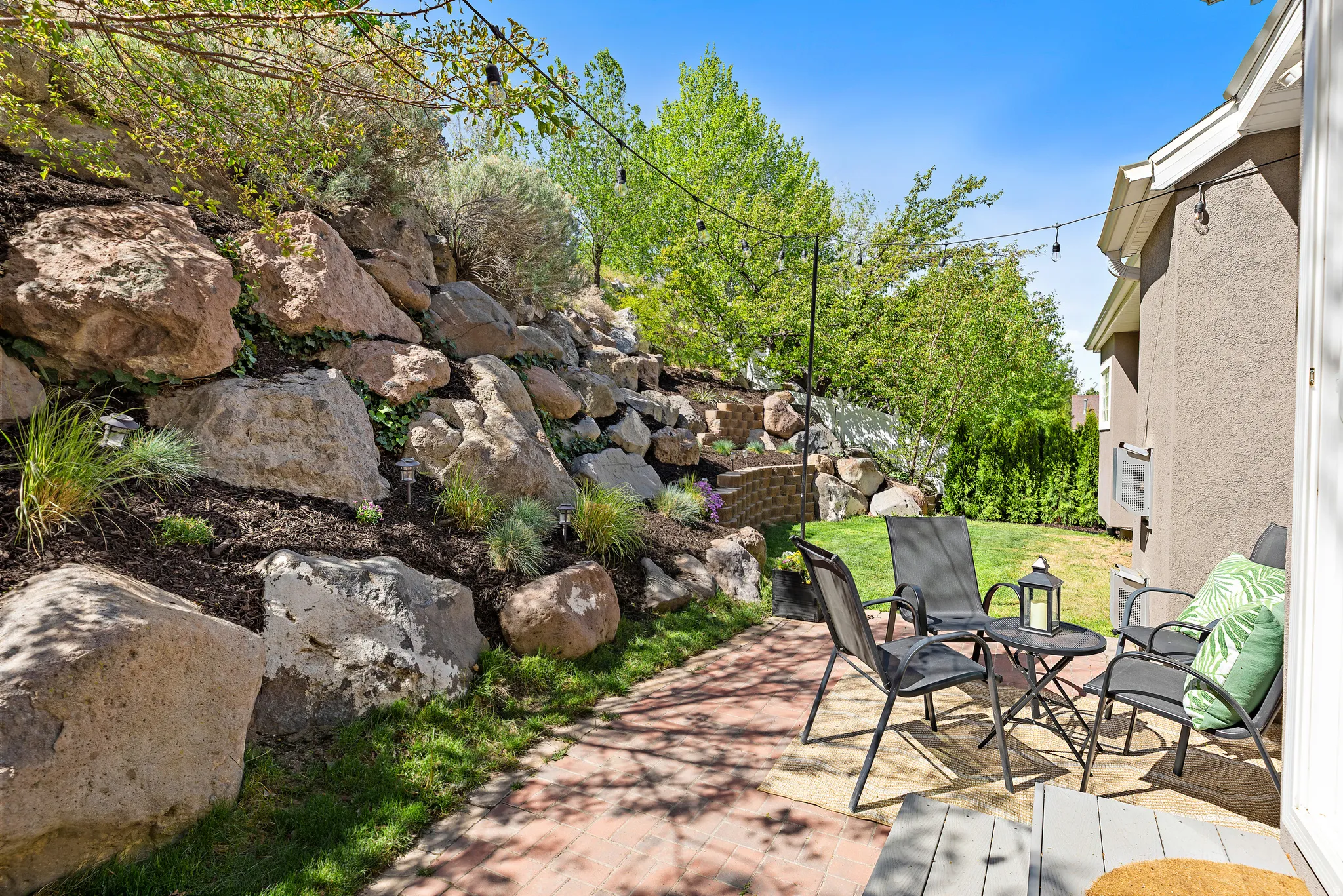Brick paver patio with a rock retaining wall, string lighting, and landscaping