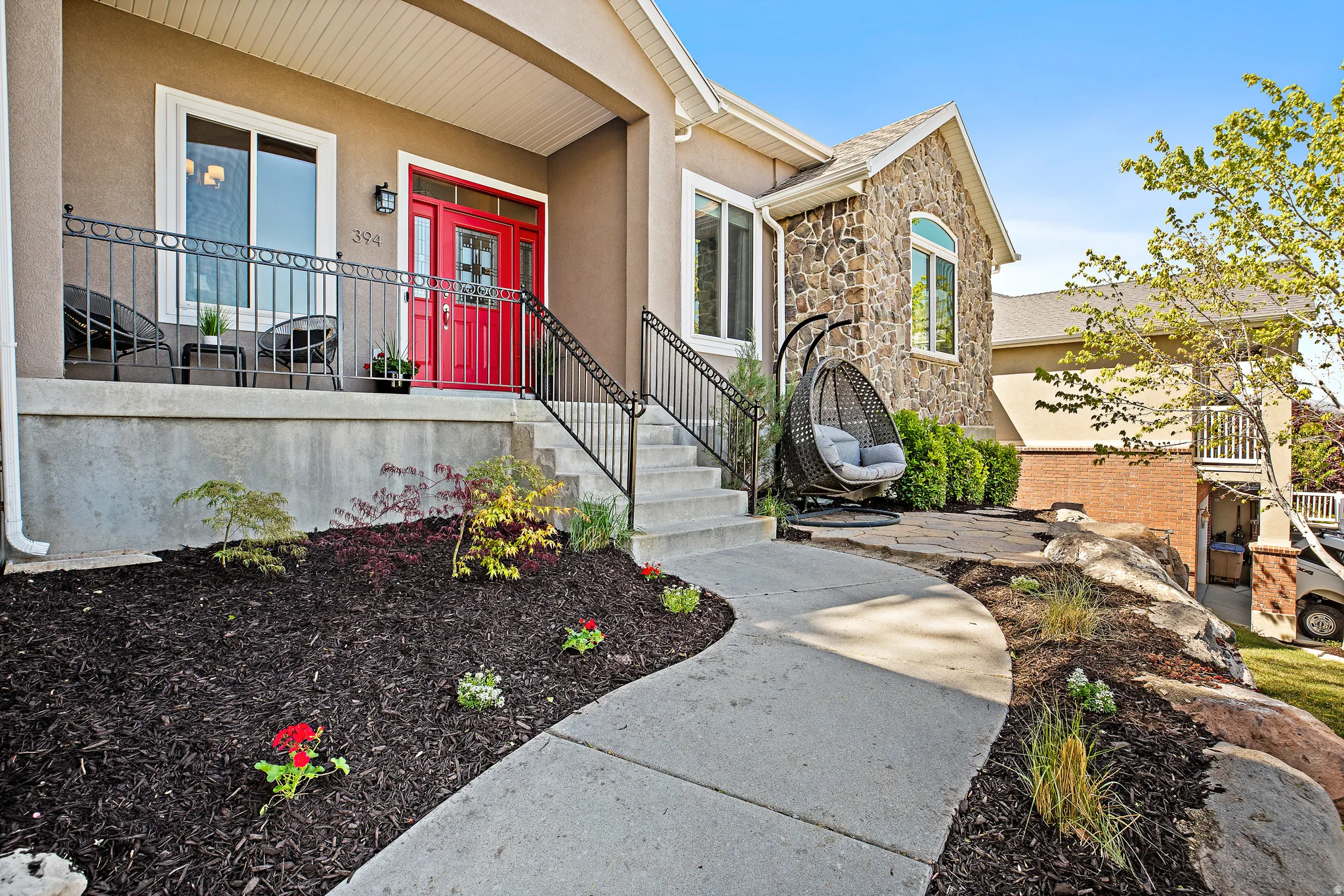 Welcoming front elevation featuring a bold red entry door, multi-level concrete walkway with metal railings, and professionally landscaped grounds with black mulch beds and seasonal plantings