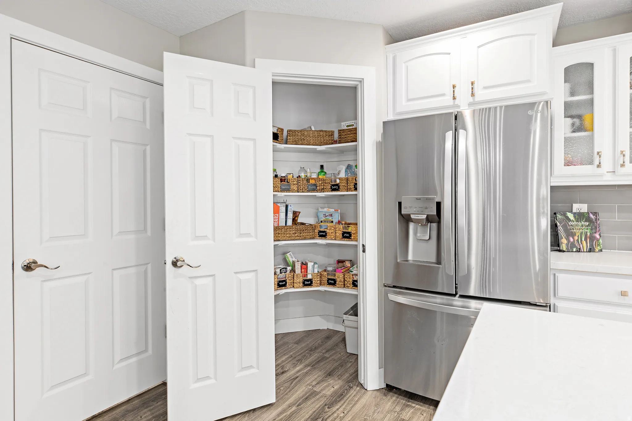 Modern kitchen featuring stainless steel French-door refrigerator with ice and water dispenser, white shaker-style cabinetry with chrome hardware, wood-finish flooring, and a large walk-in pantry with adjustable shelving and glass-front doors displaying o