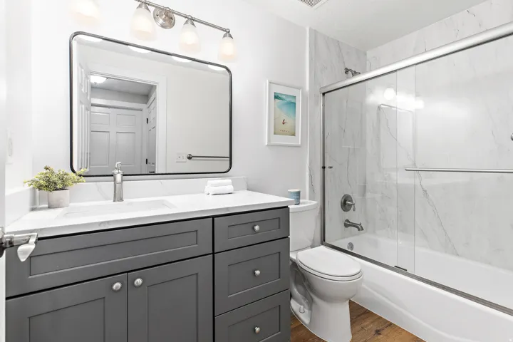 Vanity with gray cabinetry and white countertop, undermount sink with brushed nickel faucet, large framed mirror, and a shower-tub combination with glass doors and tiled surround