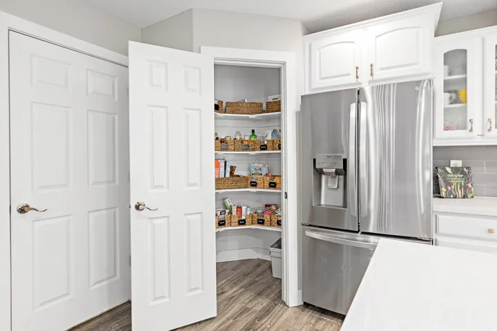Modern kitchen featuring stainless steel French-door refrigerator with ice and water dispenser, white shaker-style cabinetry with chrome hardware, wood-finish flooring, and a large walk-in pantry with adjustable shelving and glass-front doors displaying o