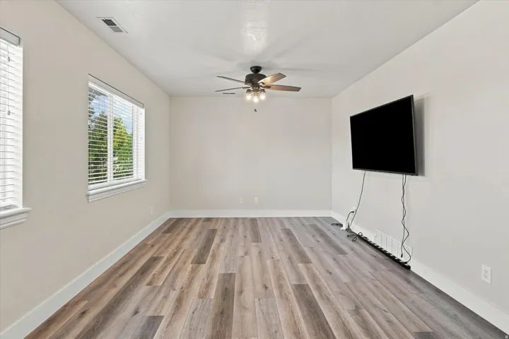 Wood-finish flooring, multiple windows with blinds, ceiling fan with integrated lighting, and a wall-mounted television