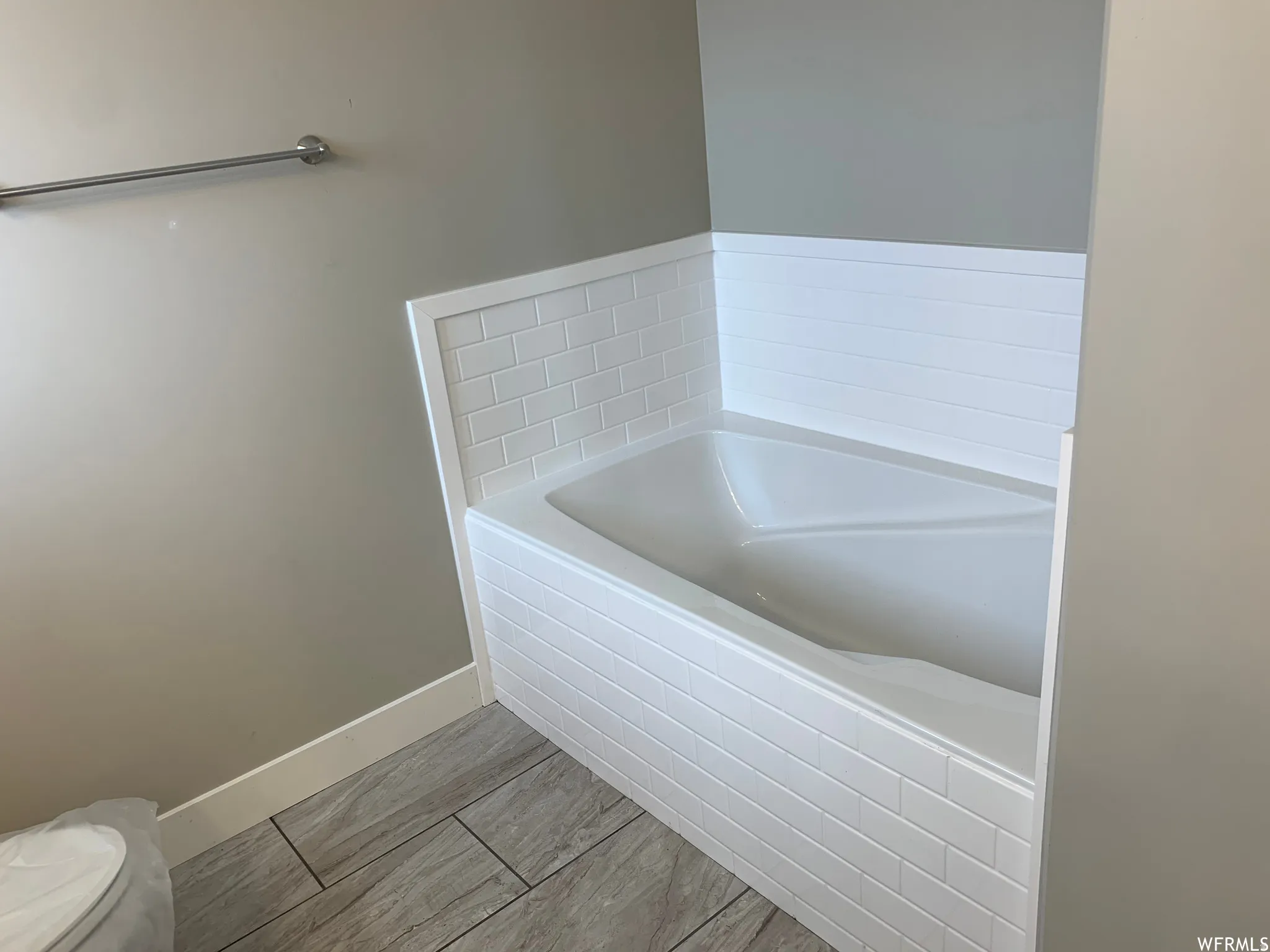 Corner soaking tub with subway tile surround, wood-finish tile flooring, and a chrome towel bar