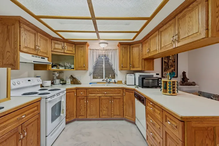 Spacious kitchen featuring warm wood cabinetry throughout, white appliances including a range and microwave, light-colored countertops, a center island with storage, coffered ceiling with skylight, and a window overlooking the grounds