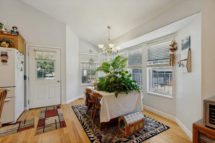 Bright, sun-filled kitchen and dining area showcasing wood-finish flooring, multiple large windows with plantation shutters, and a decorative chandelier