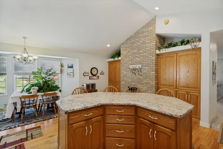 Chef's kitchen showcases custom wood cabinetry with ample storage, a generous curved island with light stone countertops and prep space, hardwood flooring, exposed brick accent wall with decorative shelving, and recessed lighting throughout