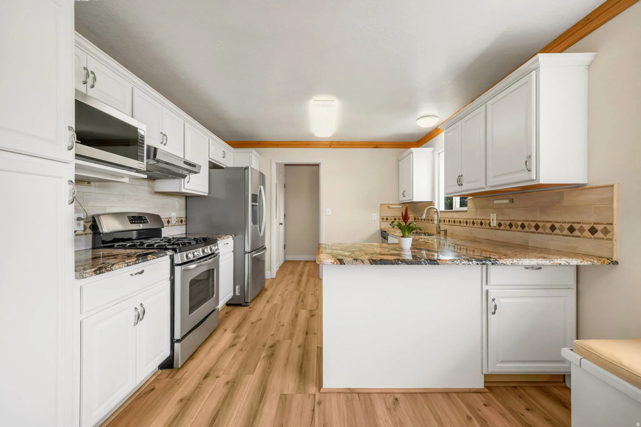 Galley-style kitchen featuring white cabinetry, stainless steel appliances, granite countertops with decorative tile backsplash, wood-finish flooring, and breakfast bar with additional counter space and storage