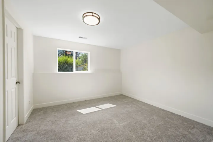 3rd basement bedroom, Recessed window with exterior greenery, flush-mount ceiling light, white panel door with chrome hardware, and New gray carpeting