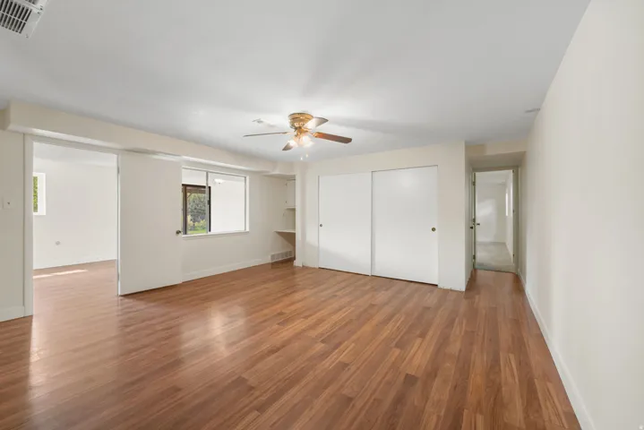 Spacious primary Bedroom/living area showcasing gleaming wood-finish flooring throughout, abundant natural light from multiple windows, and generous ceiling height enhanced by a ceiling fan with brushed brass finish