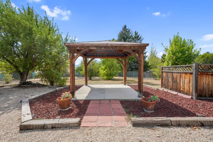 Outdoor entertaining space featuring a wood-beam pergola with a concrete foundation, brick paver walkway, and decorative red rock landscaping