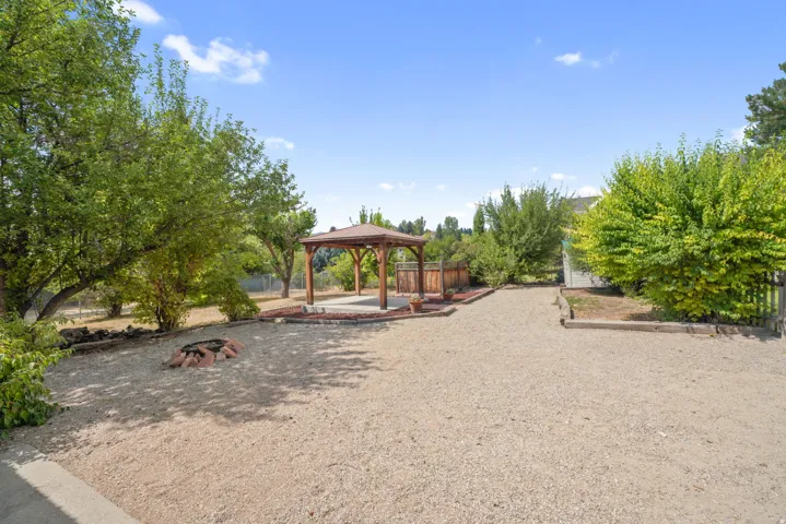 Spacious gravel yard featuring a wooden gazebo with a shingled roof, a brick-lined fire pit, and mature Fruit trees