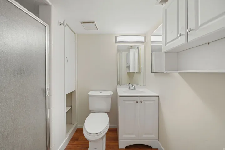 Bright bathroom featuring a white vanity with basin, Shower and ceramic toilet, recessed ceiling lighting, and wood-finish flooring throughout