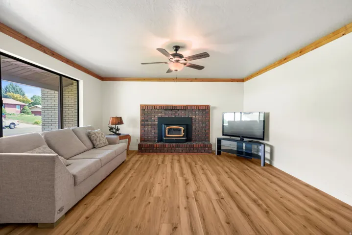 Spacious living room featuring warm wood-finish flooring throughout, a brick fireplace with ia wood burning insert, ceiling fan with integrated lighting, and large windows providing natural light
