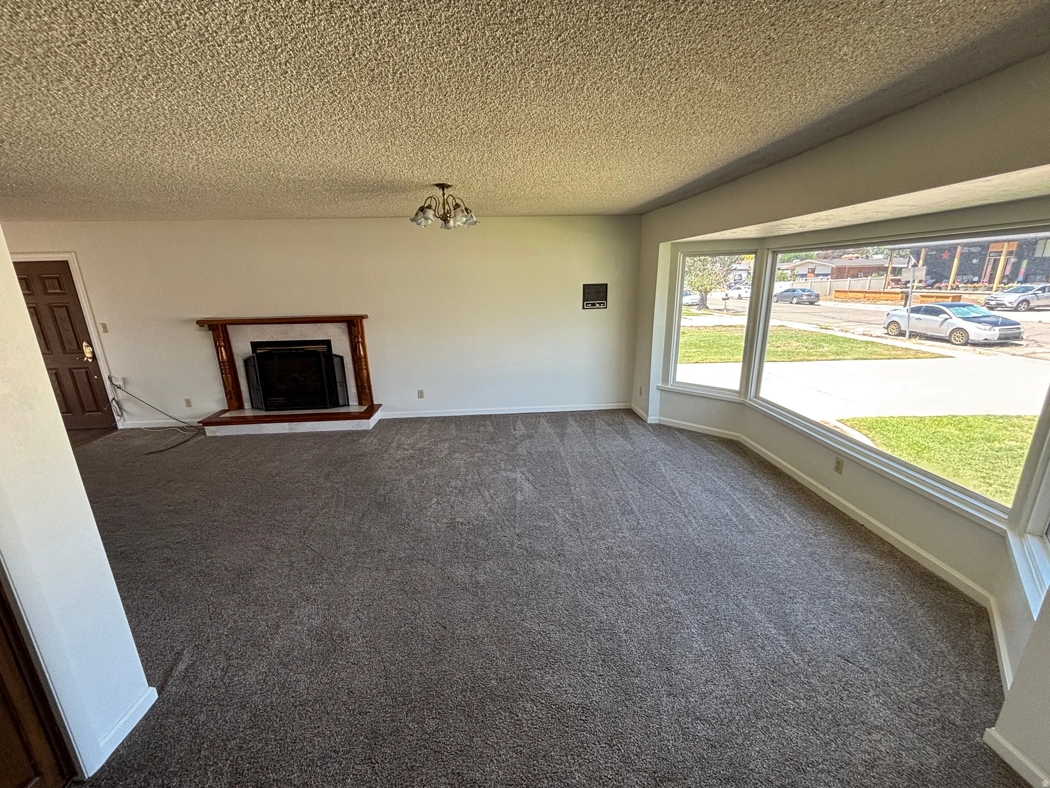 Spacious living room with neutral carpeting, textured ceiling, and abundant natural light from large picture windows overlooking the front grounds