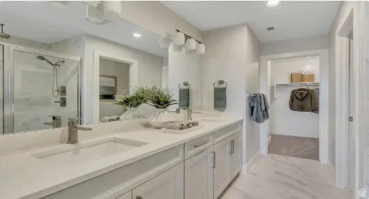 Dual sinks with brushed nickel fixtures, white cabinetry, a glass-enclosed shower, and a walk-in closet. This home is under construction. Pictures are of a similar home.