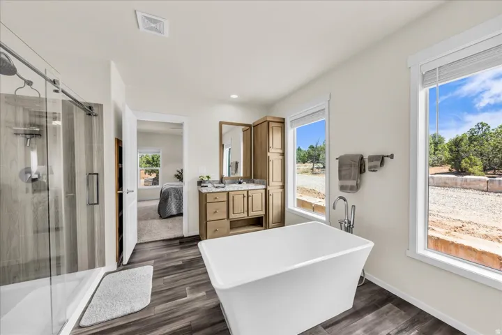 Freestanding soaking tub, dual-sink vanity with stone-finish countertop, walk-in shower with glass enclosure, wood-finish flooring, and large windows