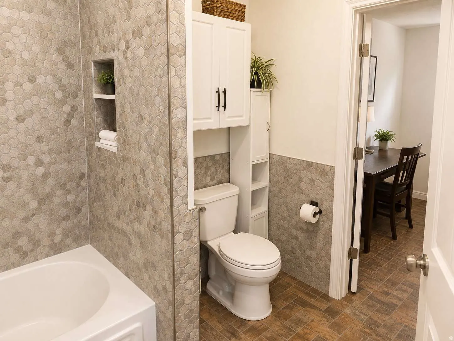 White bathtub with hexagon-tiled walls featuring a recessed niche, white toilet, and built-in white cabinetry with black hardware