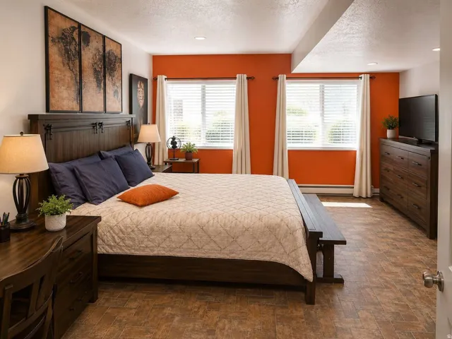 Warm-toned flooring extending throughout, two windows with blinds, a prominent headboard, and recessed lighting