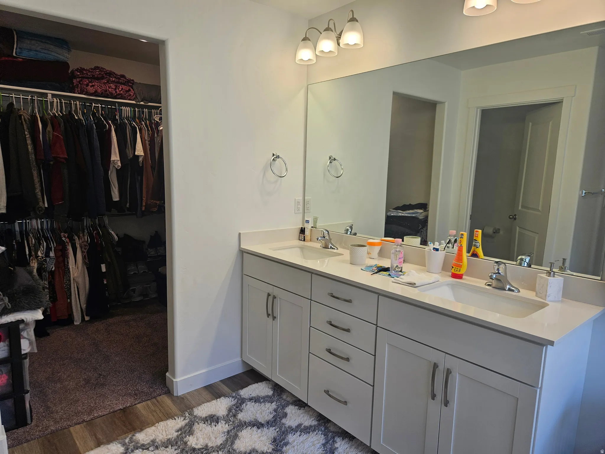 Dual vanity with integrated sinks, recessed lighting, expansive wall mirror, wood-finish flooring, and a walk-in closet