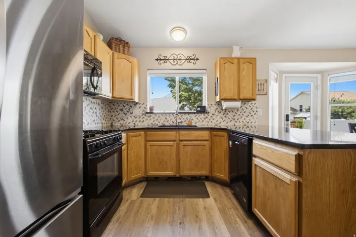 Bright kitchen featuring warm wood cabinetry, stainless steel appliances, mosaic tile backsplash, wood-finish flooring, and abundant natural light from multiple windows overlooking the surrounding landscape