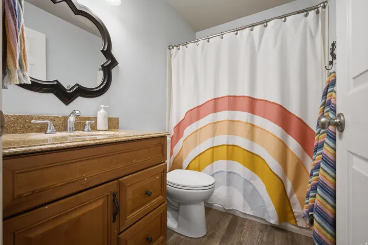 Full bathroom featuring wood-finish vanity cabinetry with a travertine countertop, white ceramic toilet, decorative mirror with ornamental black frame, and vibrant patterned shower curtain