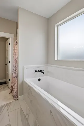 Soaking tub with a marbled surround, matte black fixtures, and a frosted window